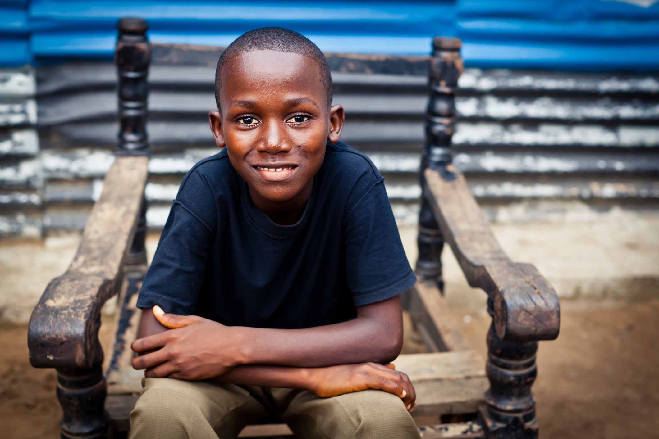 An African boy sitting in a broken chair with his hands on his knees while smiling at the camera.